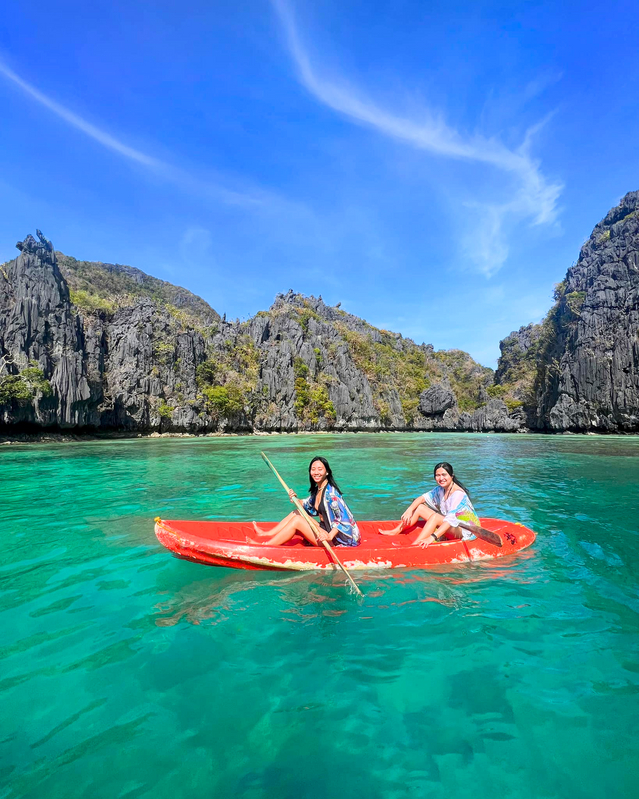 Two women kayak in a red boat on turquoise water surrounded by rocky islands under a blue sky.