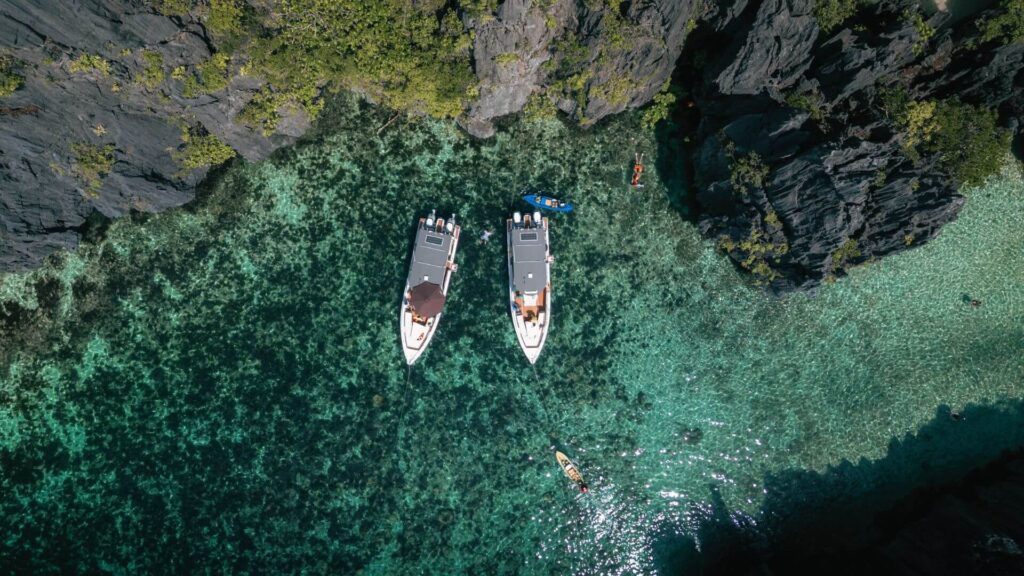 Aerial view of two boats in a clear, turquoise lagoon surrounded by rocky cliffs and kayakers.