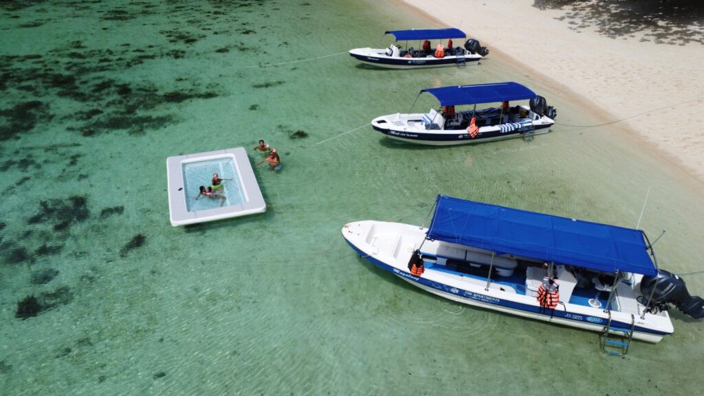 Aerial view of boats and people swimming in a floating pool in clear, shallow turquoise water near a sandy beach.