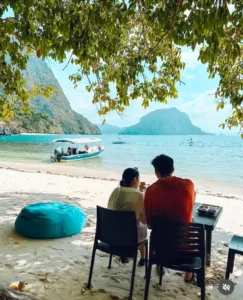Couple enjoying the beach view from a shaded table with a boat nearby.