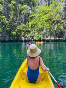 A woman in a swimsuit kayaking through turquoise waters near a rocky, tree-covered cliff.