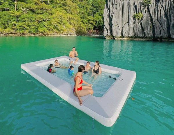 People in an inflatable pool in turquoise water with tropical greenery and cliffs in the background.