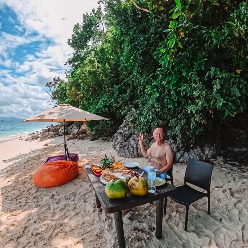 Man giving a peace sign sits at a table laden with food on a tropical beach.