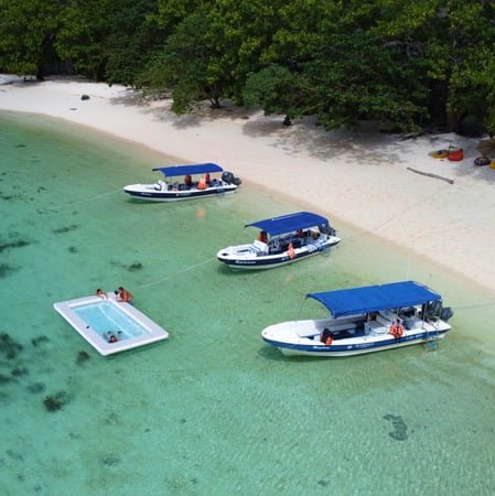 Aerial view of boats and a floating swimming pool near a sandy beach.